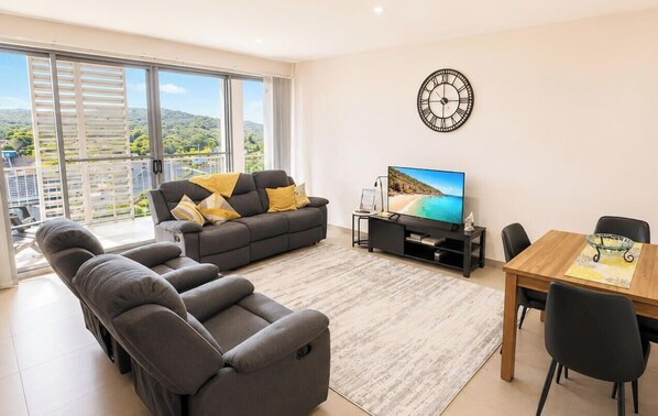 A living room with a sofa, a TV, a dining table, and a balcony with a view. - Caves Beach Retreat - close to beach and shops (Caves Beach)