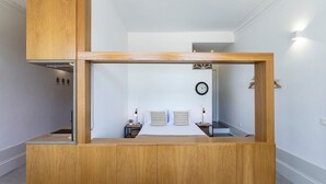 A room with a wooden cabinet, a bed with striped pillows, a bedside table, a clock, and a wall-mounted light fixture.