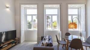 A modern living room with a flat-screen TV, a sofa, a glass dining table, and two chairs. There are three large windows with white frames and a view of palm trees and buildings outside.
