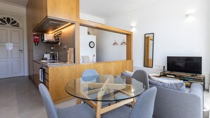 A modern living room with a glass dining table, grey sofas, a flat-screen TV, and a kitchen area with wooden cabinets and a built-in oven.