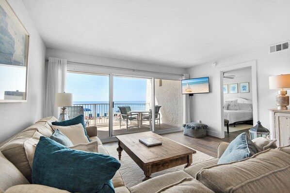 A spacious living room with a large sofa, a coffee table, and a balcony with a view of the beach.