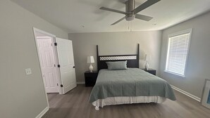 Quarto com cama, ventoinha no teto, mesa de cabeceira, abajur, janela com persianas e porta.