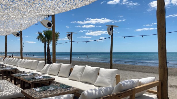Ein Sitzbereich direkt am Strand mit weißen, gepolsterten Sofas und Holztischen, mit Blick auf das Meer.
