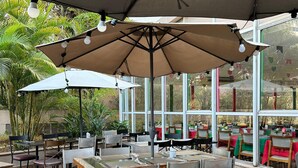 Een terras met tafels en stoelen onder parasols, omringd door grote ramen en groen.