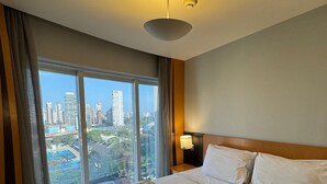 Hotelkamer met een groot bed, een nachtkastje, een lamp en uitzicht op een stadsbeeld door grote ramen.