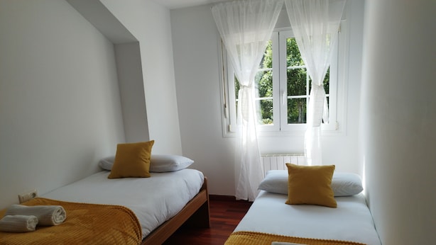 Ein Schlafzimmer mit zwei Einzelbetten, jeweils mit einer braunen Decke und einem weißen Kopfkissen. Ein Fenster mit durchsichtigen Vorhängen und ein kleiner weißer Tisch daneben.