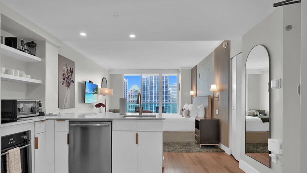A modern kitchen with white cabinetry, a dishwasher, a microwave, and a stove. There is a large window providing a view of the city skyline. - The Harmony - Ocean & Relaxation (Miami)