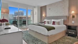 A modern hotel room with a large bed, a desk with a vase of flowers, a city view through the glass door, and a nightstand with a lamp. - The Harmony - Ocean & Relaxation (Miami)