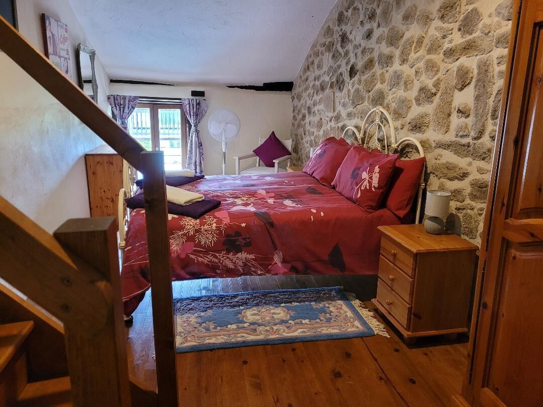 A bedroom with a stone wall, a wooden bed with red bedding, a wooden dresser, and a fan.