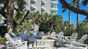 A patio area with white Adirondack chairs and a stone fire pit surrounded by lush greenery and a multi-story building in the background. - Modern 4th Floor Villa w/ Private Balcony (Hilton Head Island)