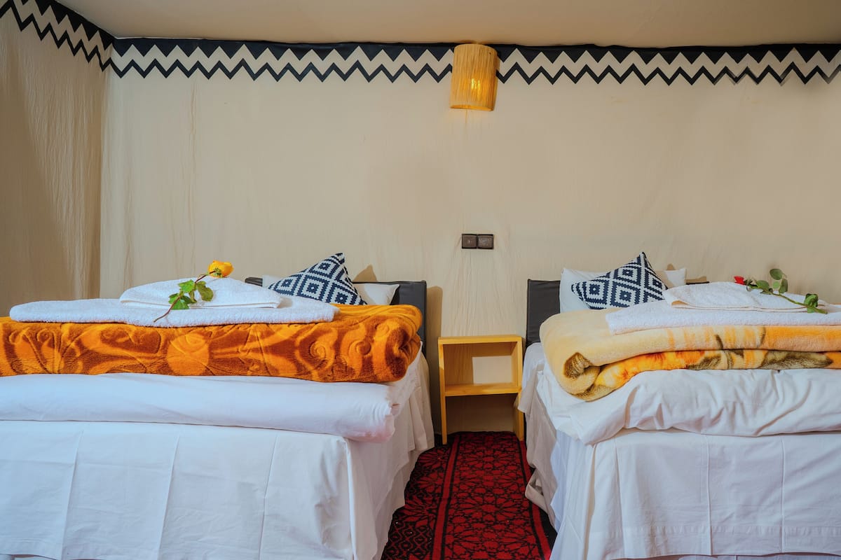 A hotel room with two single beds, each with a white duvet and a patterned pillow. There is a small wooden nightstand between the beds, and a wall-mounted light fixture above.