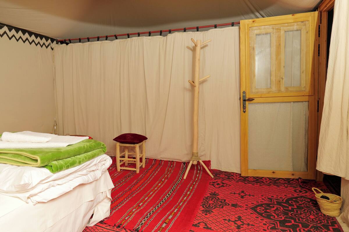 A room with a bed, a wooden coat rack, a small stool, a patterned rug, and a door with a mesh screen.