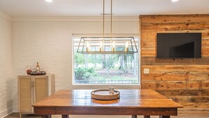 A dining area with a wooden table, a chandelier, a television mounted on a wooden wall, and a window with a view of greenery.