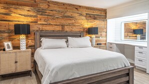 A bedroom with a wooden headboard, a bed with white bedding, two bedside tables with lamps, and a window with a view of the outside.