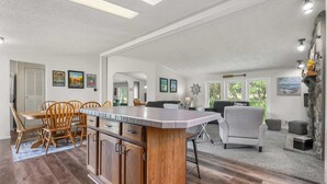 Un vaste espace de vie comprenant un îlot de cuisine, une table à manger et des chaises. Les murs sont ornés de tableaux encadrés, il y a une grande fenêtre et un mur d’accent en pierre.