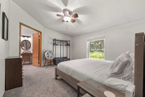 Ein Schlafzimmer mit Bett, Deckenventilator, Holzschrank, Spiegel, Stuhl und Fenster mit Blick auf Grünflächen.