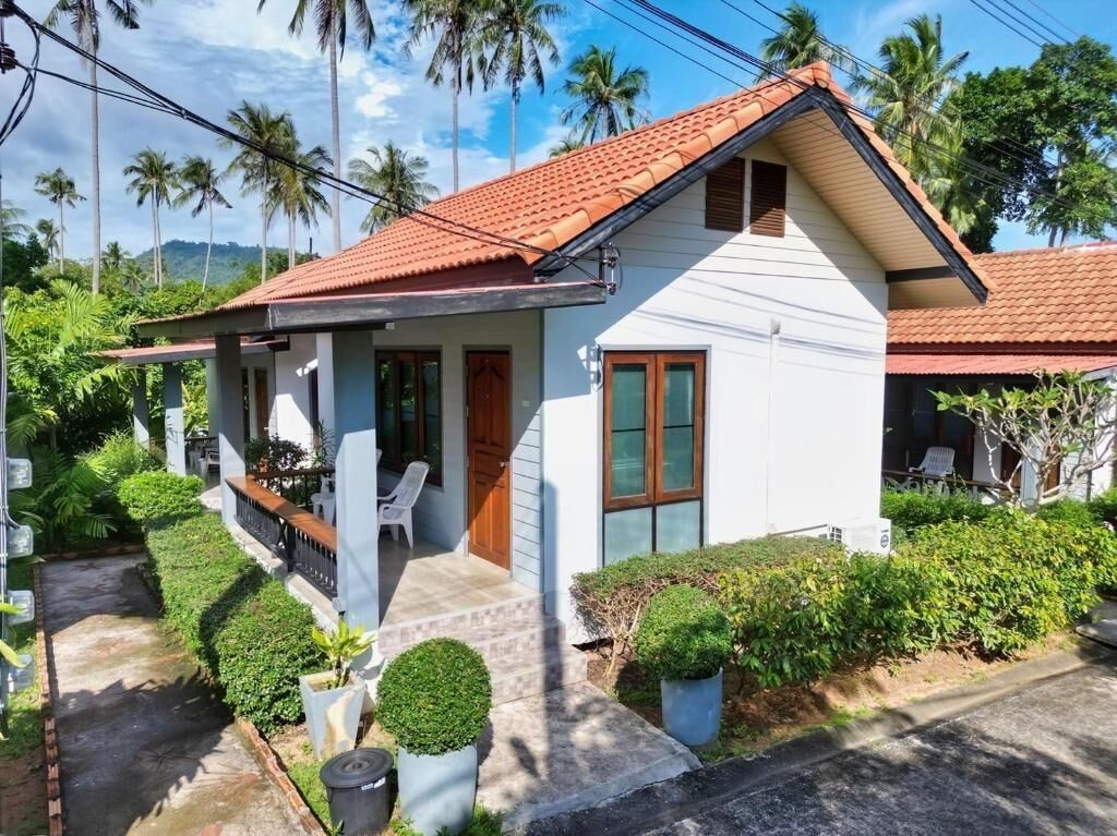 A small, single-story house with a red-tiled roof, a porch with chairs, and a well-maintained garden.