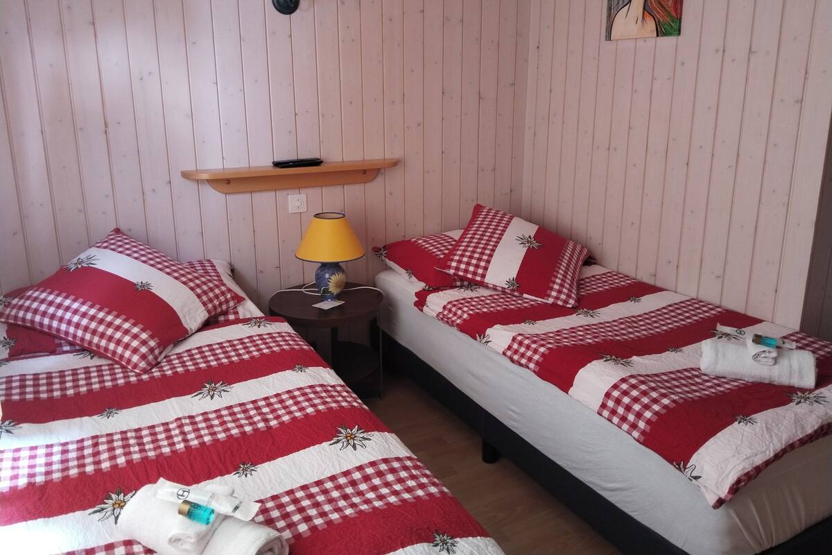 Huoneessa on kaksi sänkyä, joissa on punavalkoruudulliset vuodevaatteet ja kukkakuviot. Sänkyjen yläpuolella olevalla hyllyllä on pieni pöytä, jossa on keltainen lamppu ja kaukosäädin.