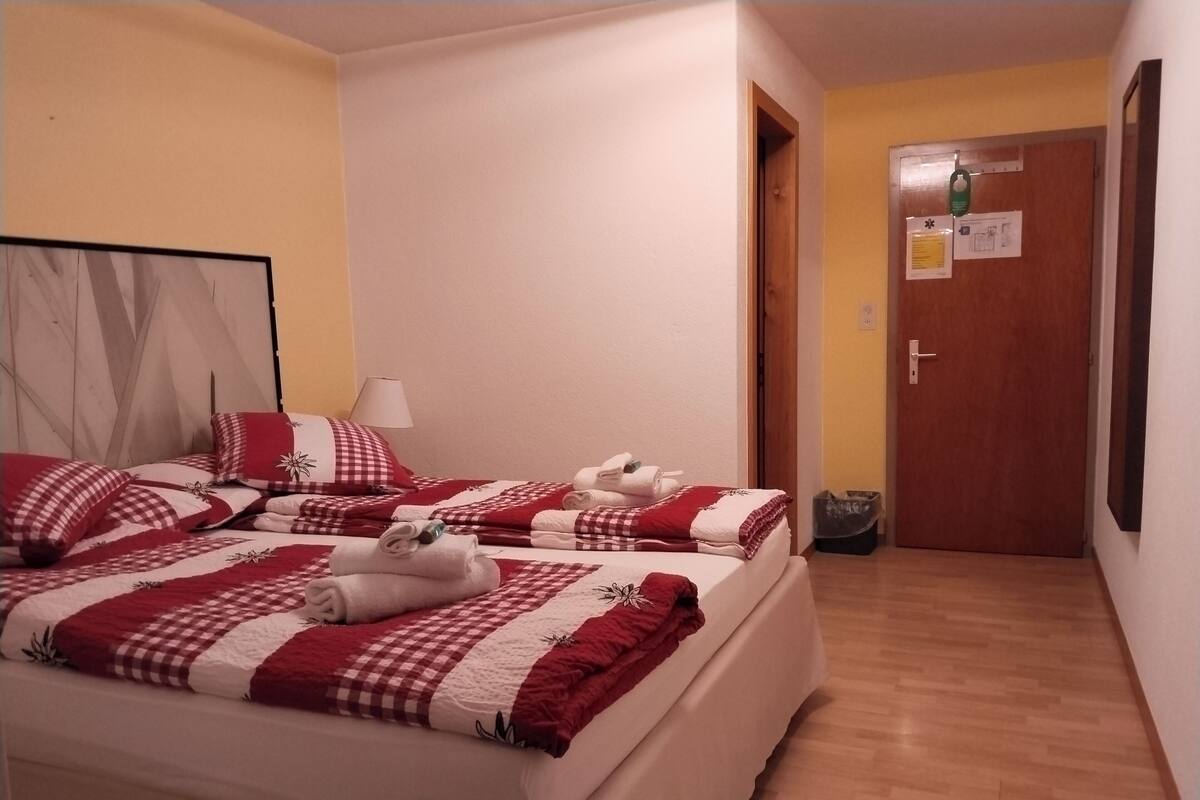 Hotellihuone, jossa on kaksi sänkyä. Kummassakin sängyssä on punavalkoruudullinen vuodevaatesetti, huoneessa on puulattia ja ovi, jossa on lappu.