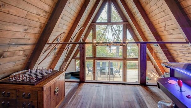 Een ruime zolderkamer met houten balken, een tafelvoetbaltafel en een groot raam met uitzicht op het landschap.