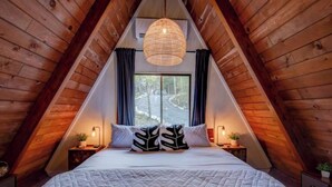 Een knus slaapkamer onder de dakpannen, met een groot bed, een hanglamp en een raam met gordijnen.