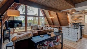 Een gezellig loft-achtig vertrek met een groot raam, houten balken in het plafond, een bank, een eettafel en een keukenhoek.
