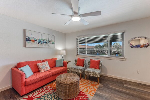 A living room with a red sofa, patterned chairs, a wicker ottoman, a ceiling fan, a window with blinds, and a wall-mounted mirror. - Beach Daze- Roomy1 BDRM a block from the BEACH & Large Pool (Port Aransas)