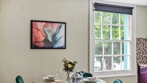 A dining area with a round glass table, green upholstered chairs, a vase with flowers, and a framed abstract painting on the wall.