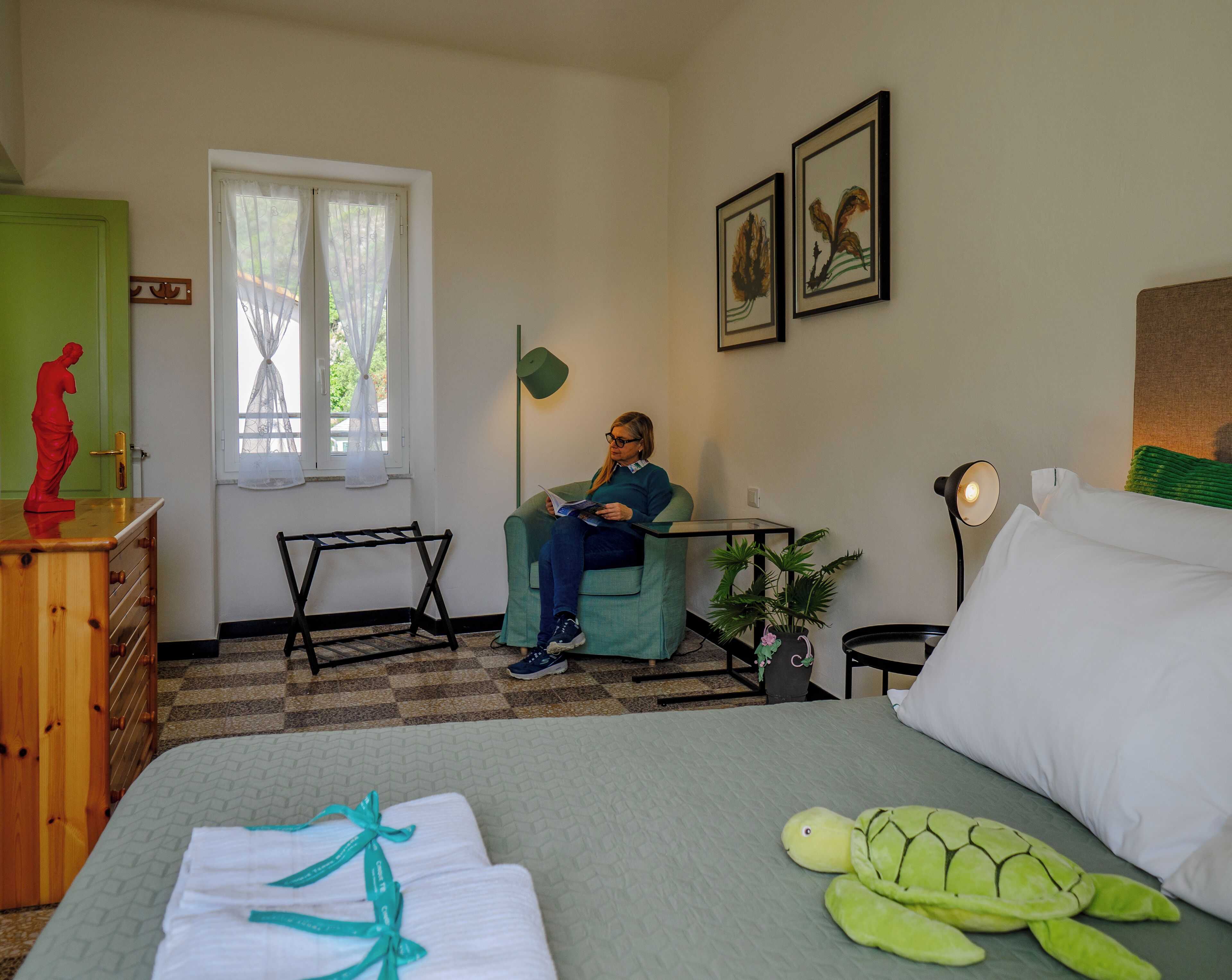 Una habitació amb un llit, una cadira, una taula, una làmpada i una finestra amb cortines.