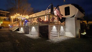 Una caravana bien iluminada con terraza cubierta, asientos exteriores y banderas estadounidenses.