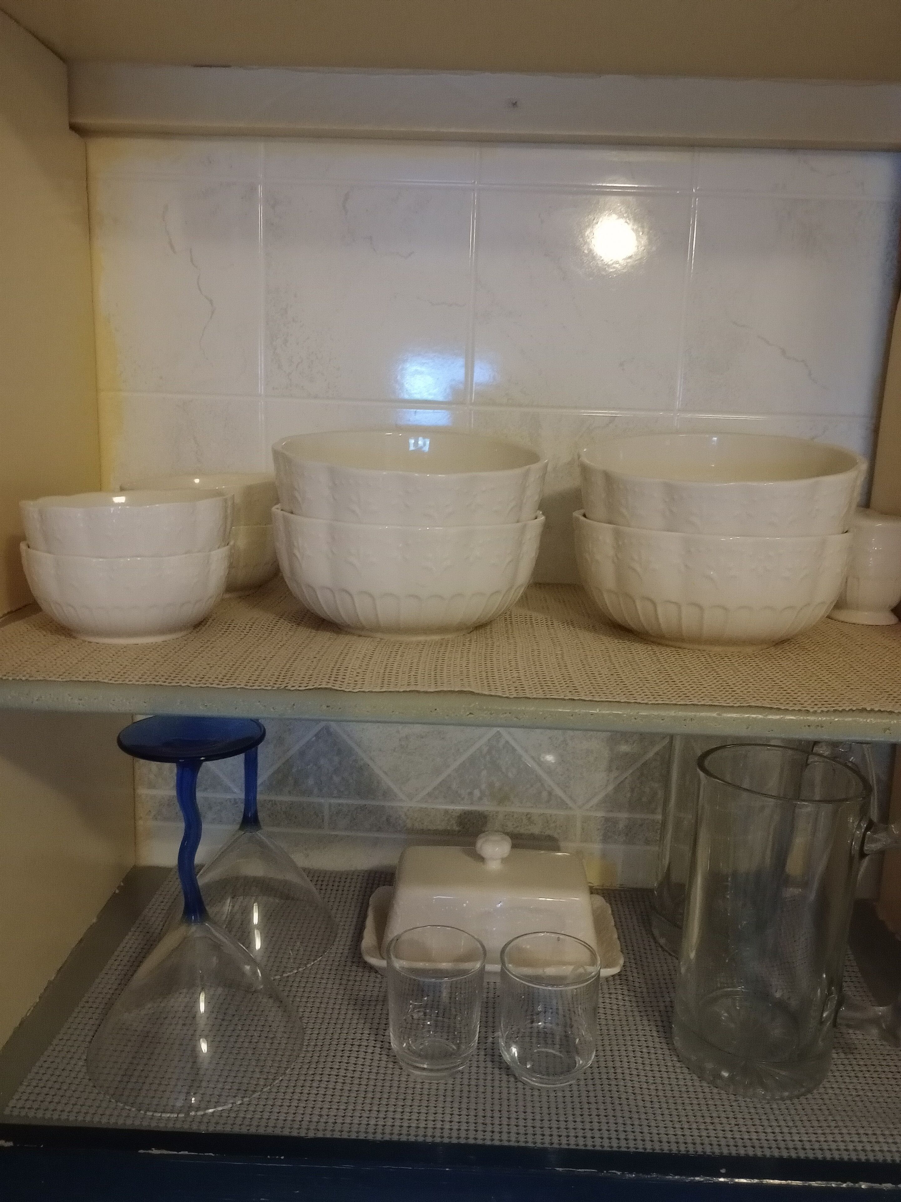 A kitchen with white ceramic bowls and cups on a shelf, a blue stool, and a glass pitcher on a tray.