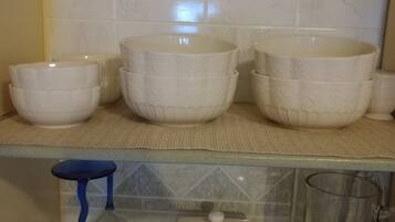 A kitchen with white ceramic bowls and cups on a shelf, a blue stool, and a glass pitcher on a tray.
