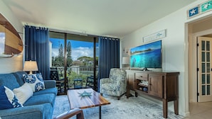 Sala de estar com sofá, poltrona, mesa de centro, televisão e vista para árvores e céu através de grandes janelas.