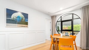 Un coin repas avec une table ronde et des chaises orange, une grande fenêtre donnant sur un paysage verdoyant et une œuvre d’art encadrée accrochée au mur.