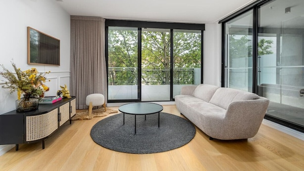 Un salon moderne comprenant un canapé, une table basse ronde, une table d’appoint avec un vase de fleurs, une console noire et une grande fenêtre donnant sur des arbres.
