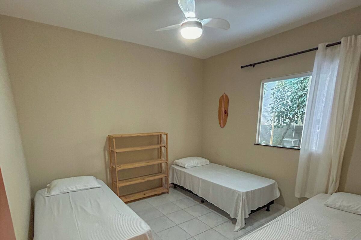 Quarto com três camas de solteiro, um ventilador de teto, uma janela com cortinas e uma prateleira de madeira.