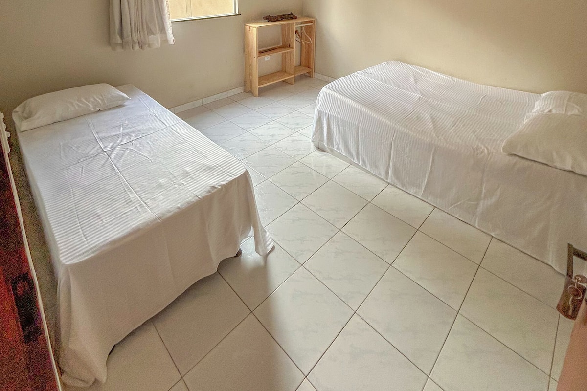 Quarto com duas camas de solteiro, um ventilador de teto, janela com cortinas, uma pequena mesa de madeira e um adorno na parede.