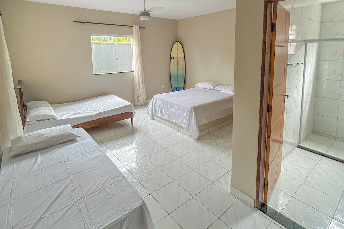 Quarto com duas camas de solteiro, ventoinha no teto, uma prancha de surf e área de chuveiro.