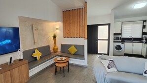 Ein modernes Wohnzimmer mit Flachbildfernseher, einer Couch, einem kleinen Tisch und einer Küchenzeile mit Geräten.