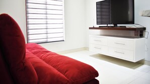 Une pièce avec un canapé rouge, un téléviseur posé sur un meuble blanc et une fenêtre avec des stores.
