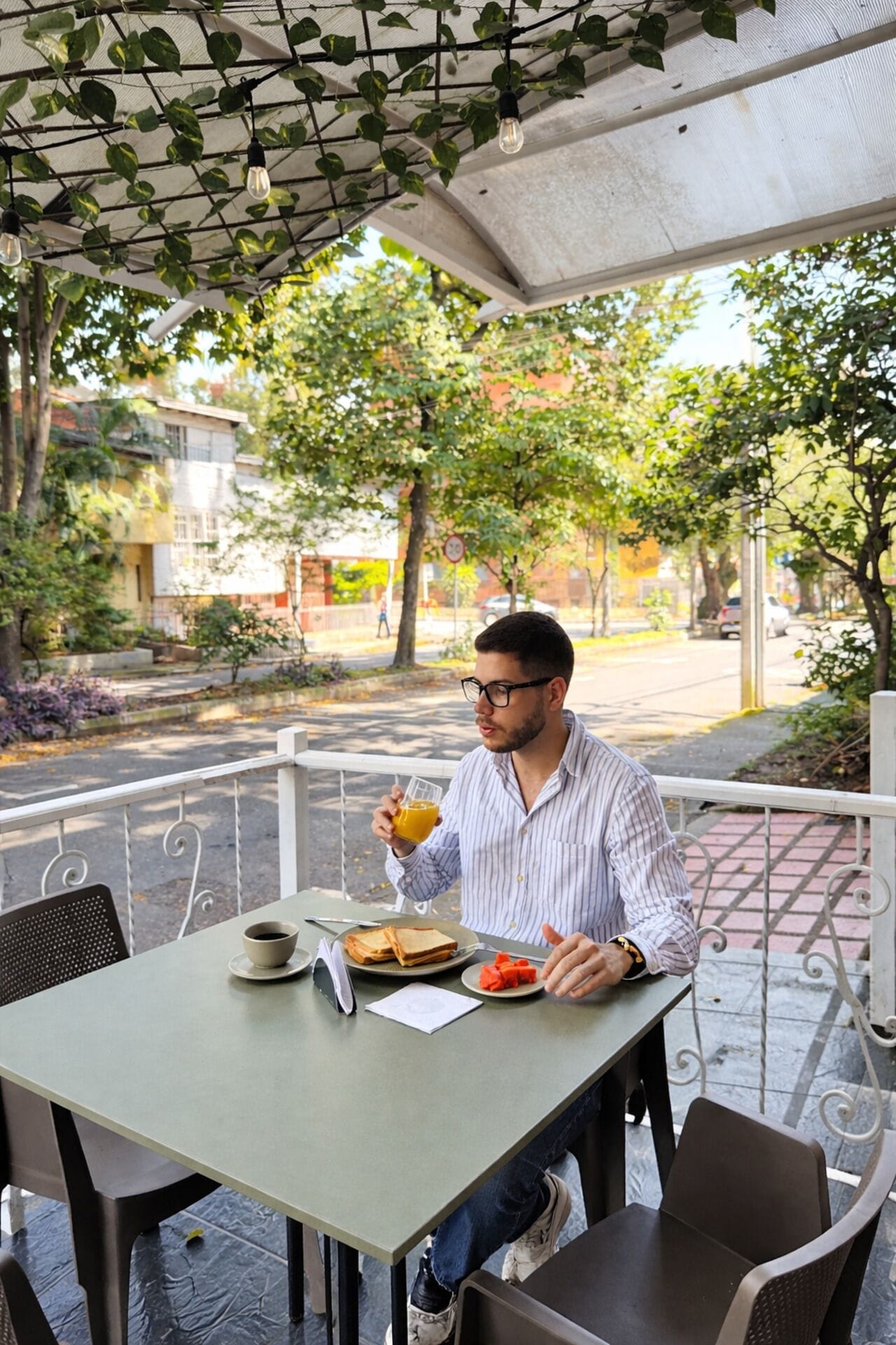 A person sitting at an outdoor table with a drink, a cup, and a plate of food.