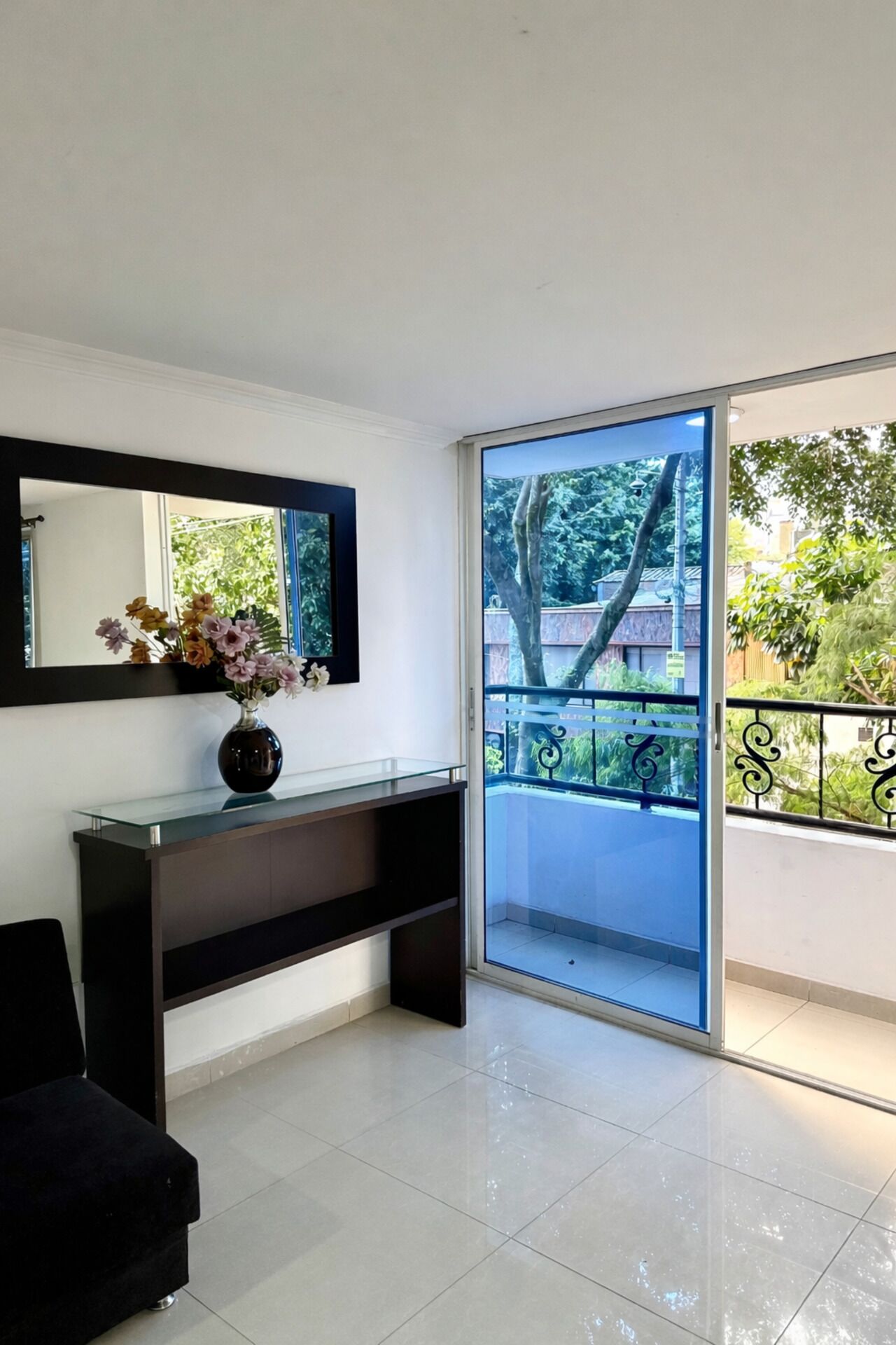 A modern living room with a glass door leading to a balcony, a dark console table with a vase of flowers, and a large mirror on the wall.