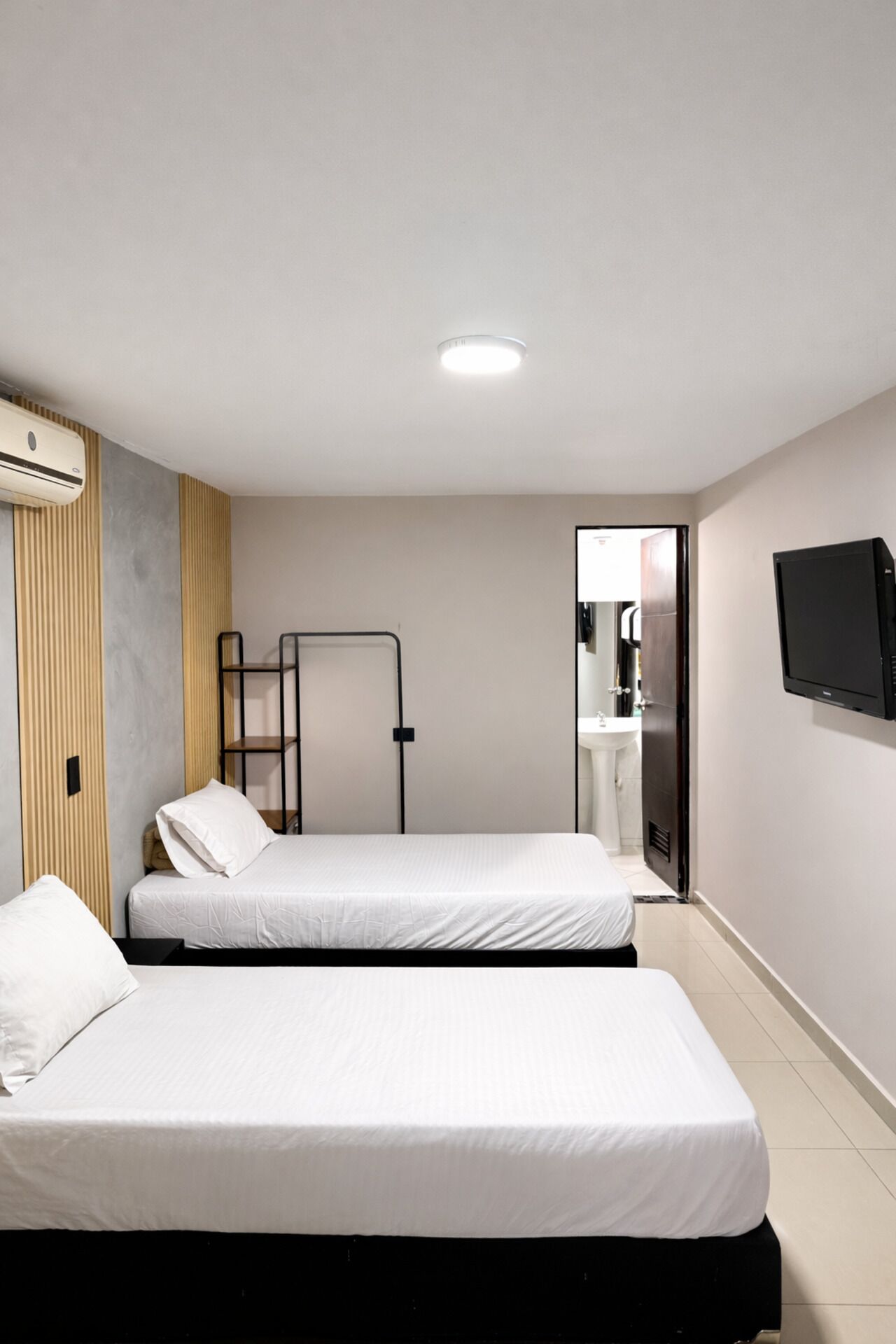 A small, modern room with two beds, a TV, and a bathroom visible through an open door.