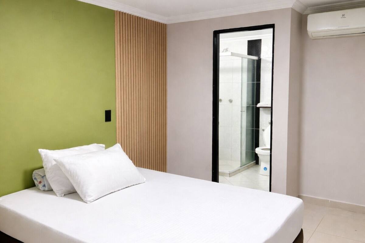 A bedroom with a bed, a green wall, a wooden panel, a bathroom with a glass door, and an air conditioning unit.