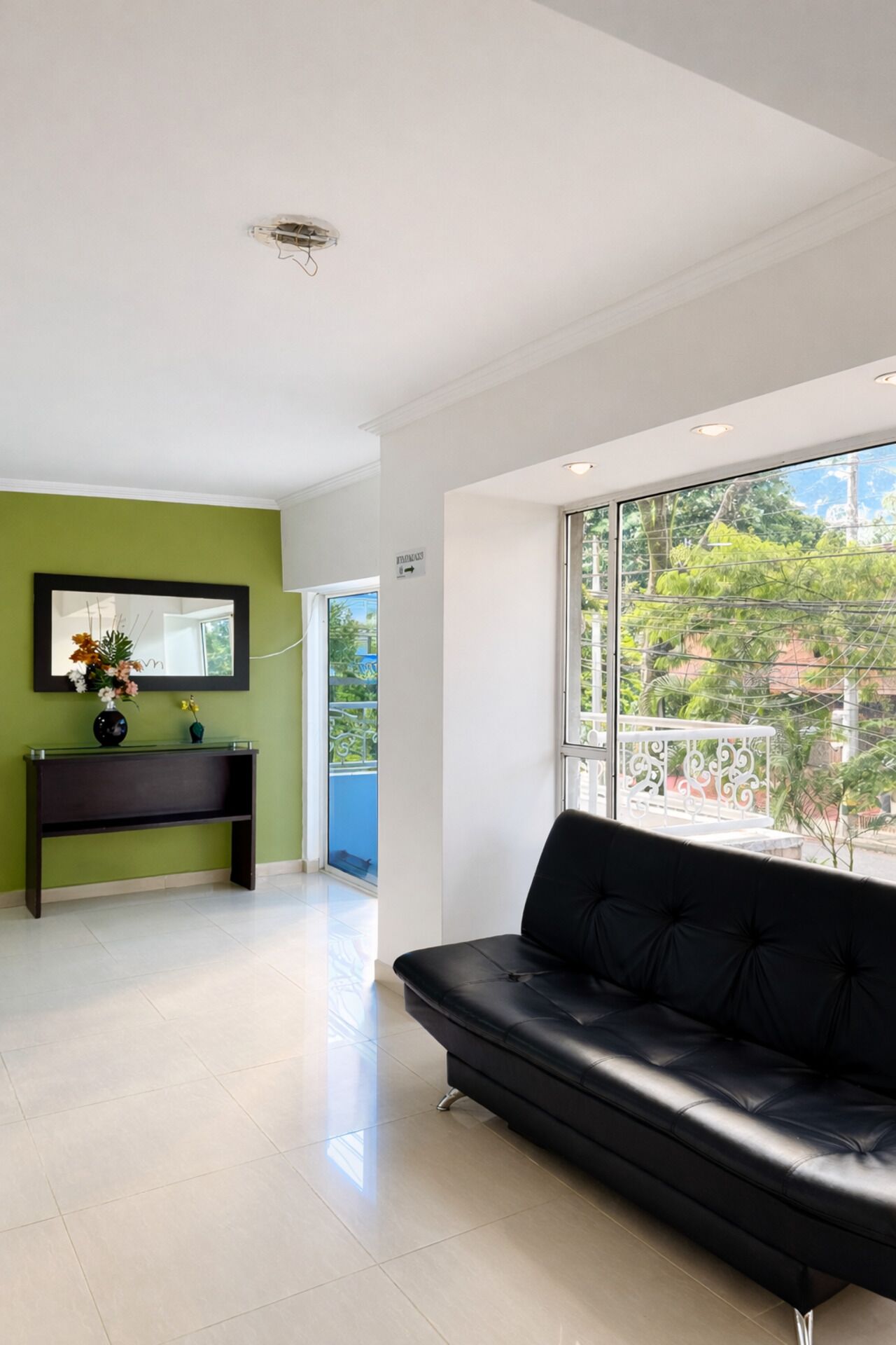 A modern living room with a black leather sofa, a glass console table with a vase, a large window with a view of greenery, and a green accent wall.