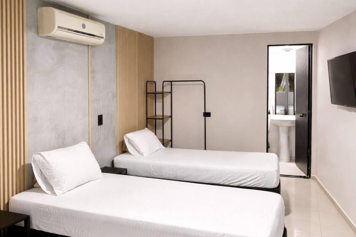 A modern hotel room with two single beds, a built-in wardrobe, a flat-screen TV, and a bathroom with a sink and mirror.