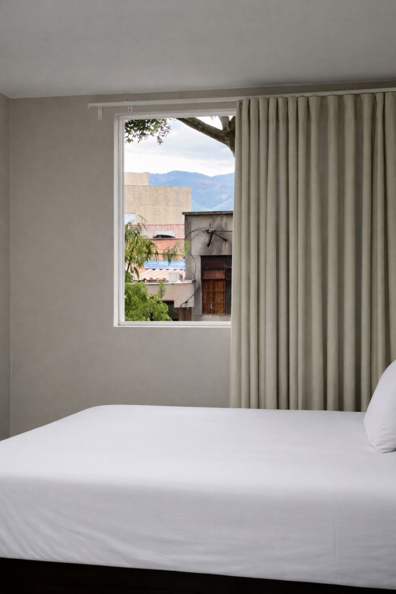 A hotel room with a bed, a window with curtains, and a view of buildings and mountains.