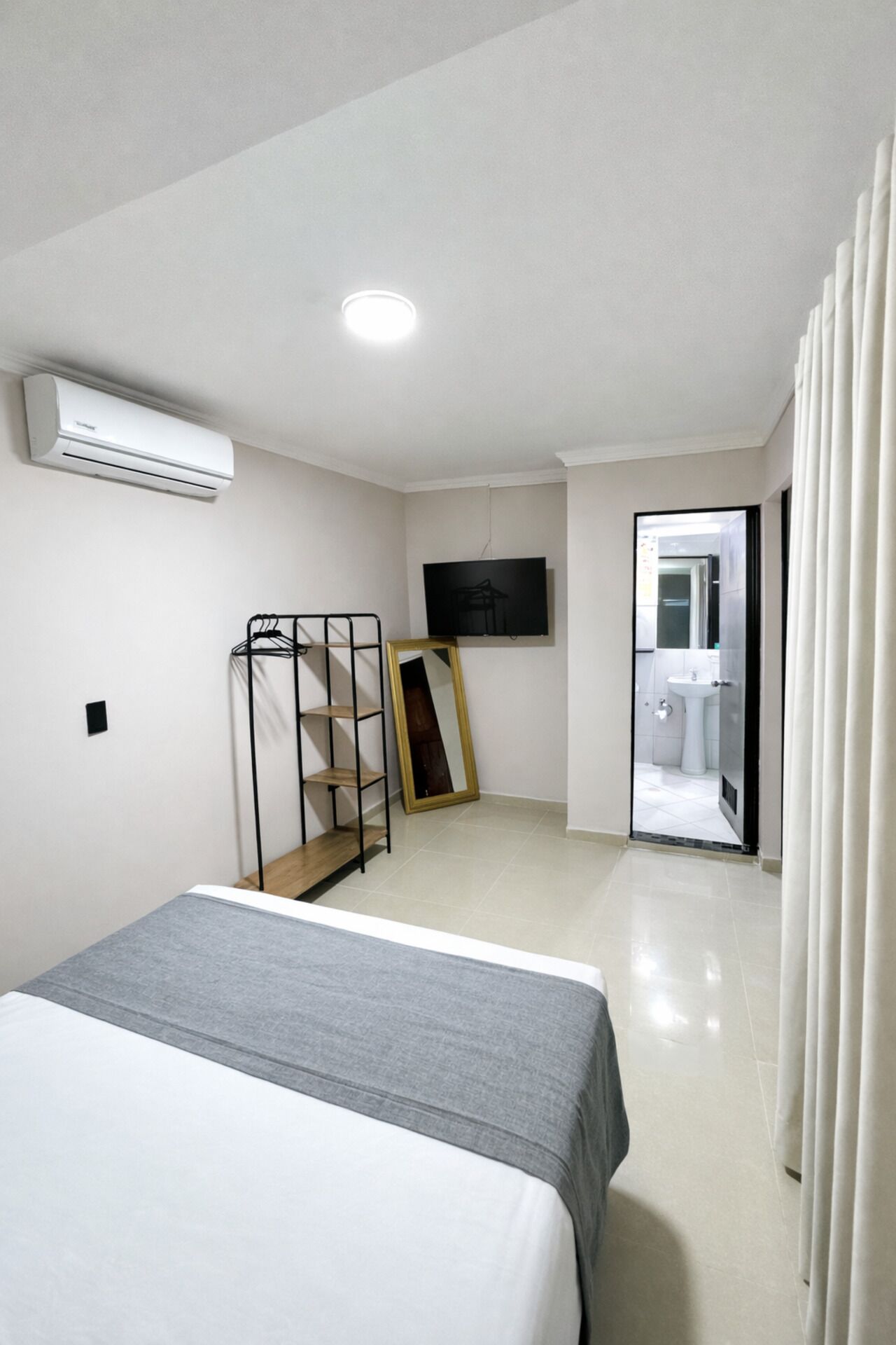 A modern hotel room with a bed, a TV, a mirror, and a bathroom visible through an open door.