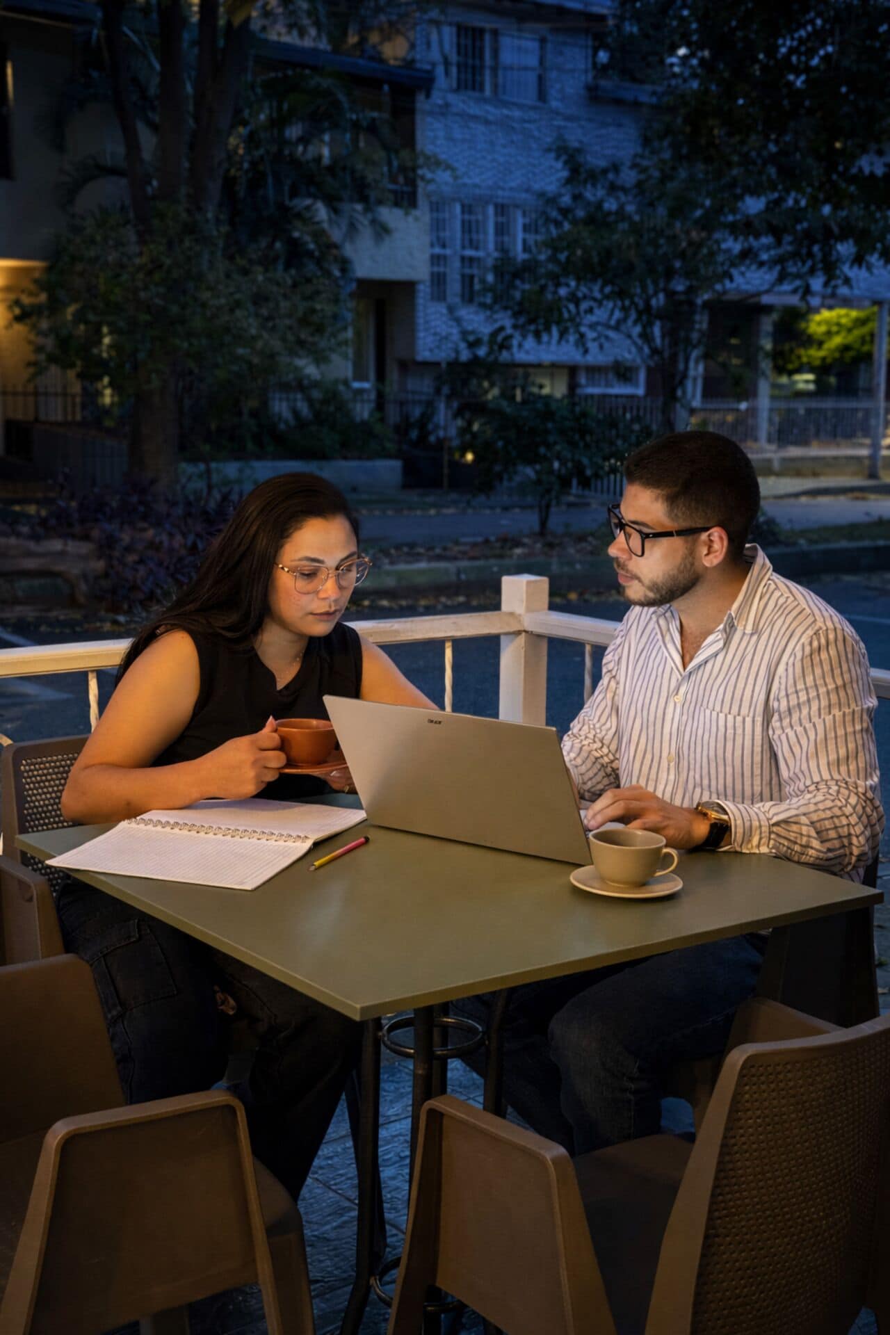Zwei Personen sitzen an einem Tisch im Freien, mit einem Laptop und einer Tasse, umgeben von Stühlen und einem Notizbuch.