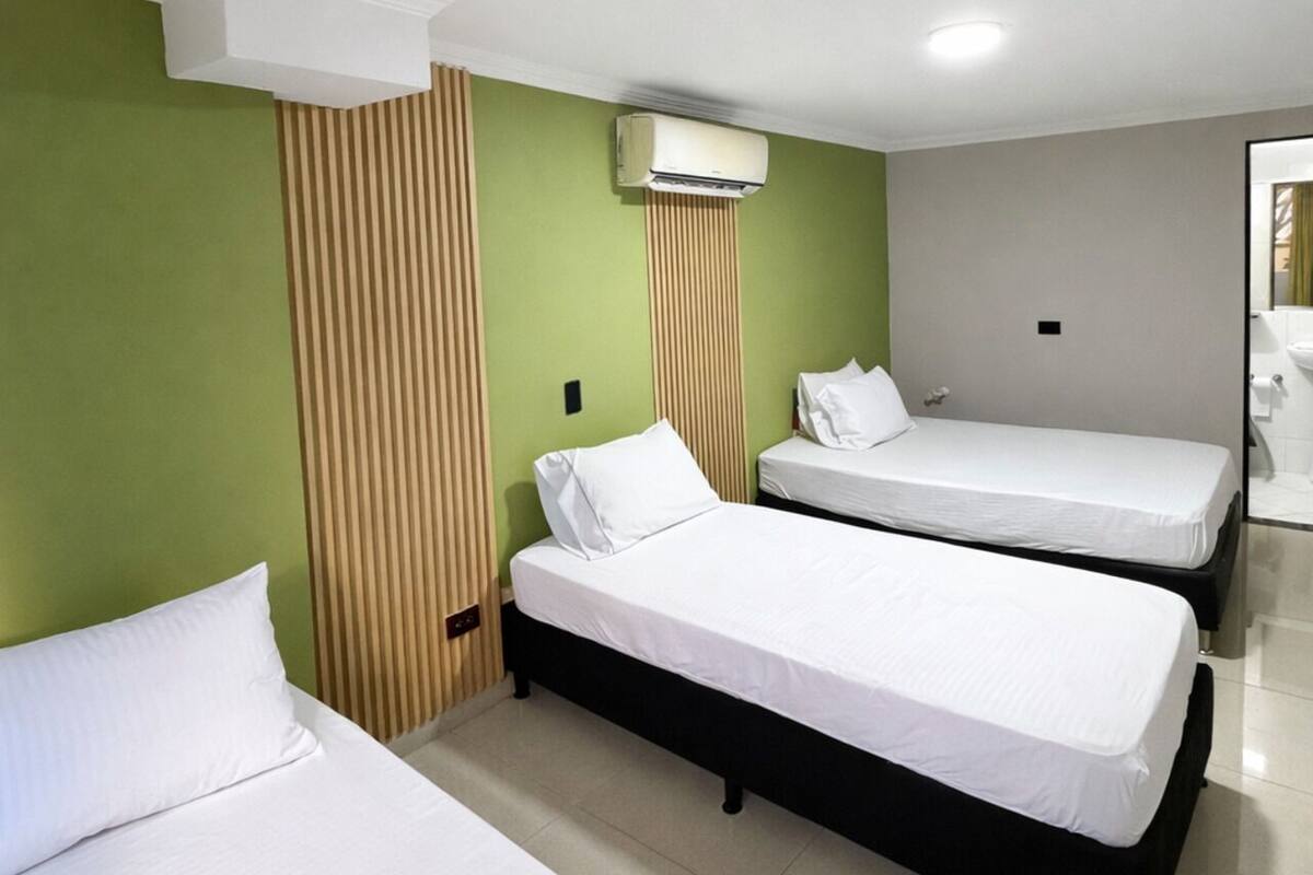 A hotel room with two beds, a window, a wall-mounted air conditioner, and a bathroom with a sink and mirror.